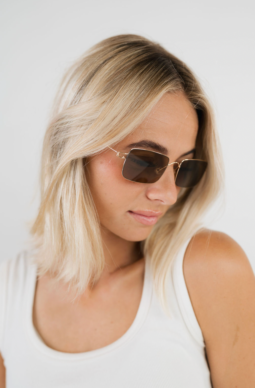 A woman with blonde hair wearing square aviator-style sunglasses with gold metal frames and dark brown lenses, paired with a white tank top against a plain white background.