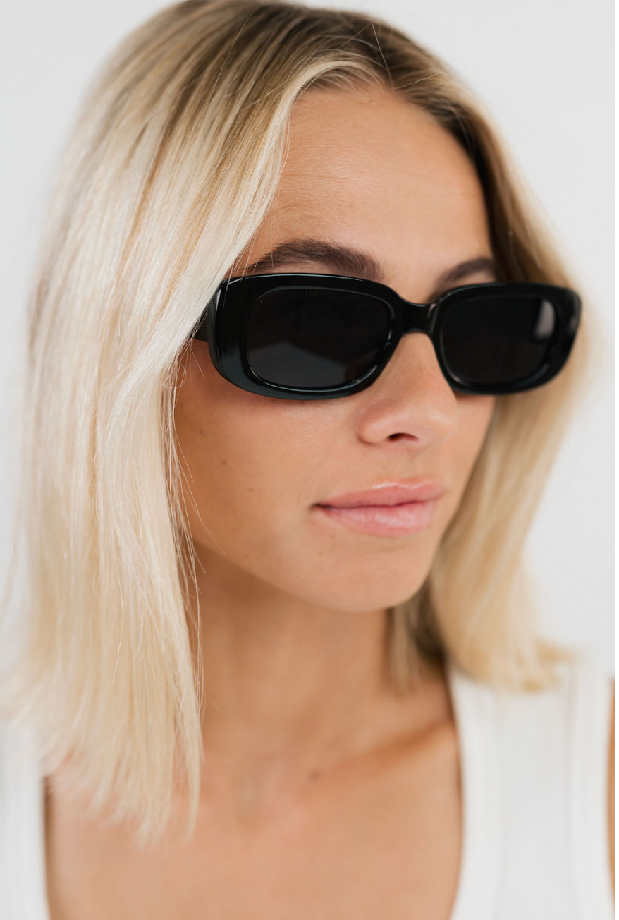 A close-up of a woman with blonde hair wearing black rectangular sunglasses and a white top against a clean white background.
