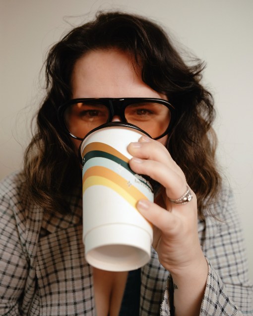 A woman in a plaid blazer drinking from a paper cup, wearing oversized black aviators with warm amber lenses