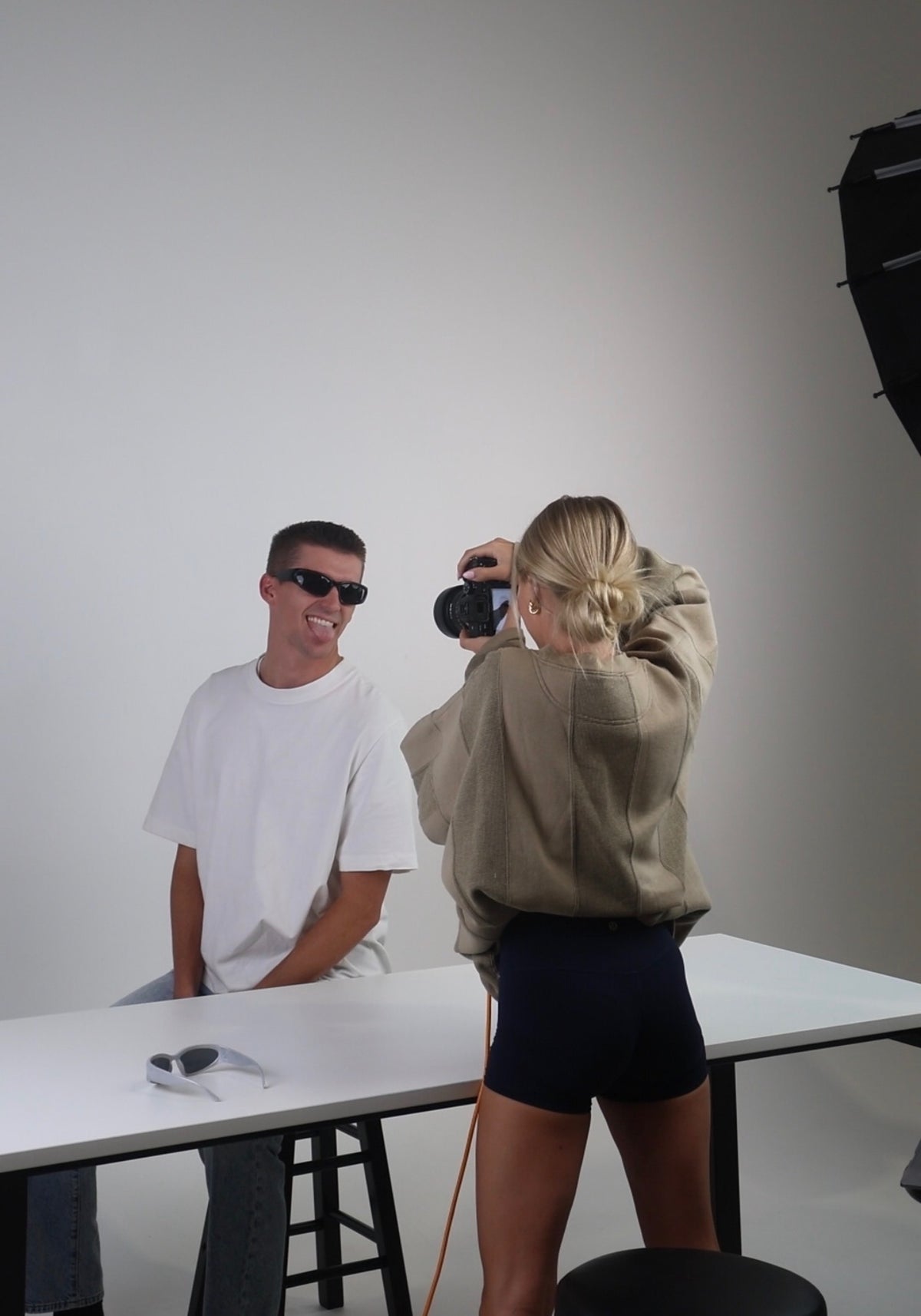 A lifestyle studio scene showing a man in a white t-shirt and sunglasses sitting on a stool, while a woman in a beige hoodie and black shorts takes a photo of him with a camera. A white table and a studio light are visible against a plain light gray background.