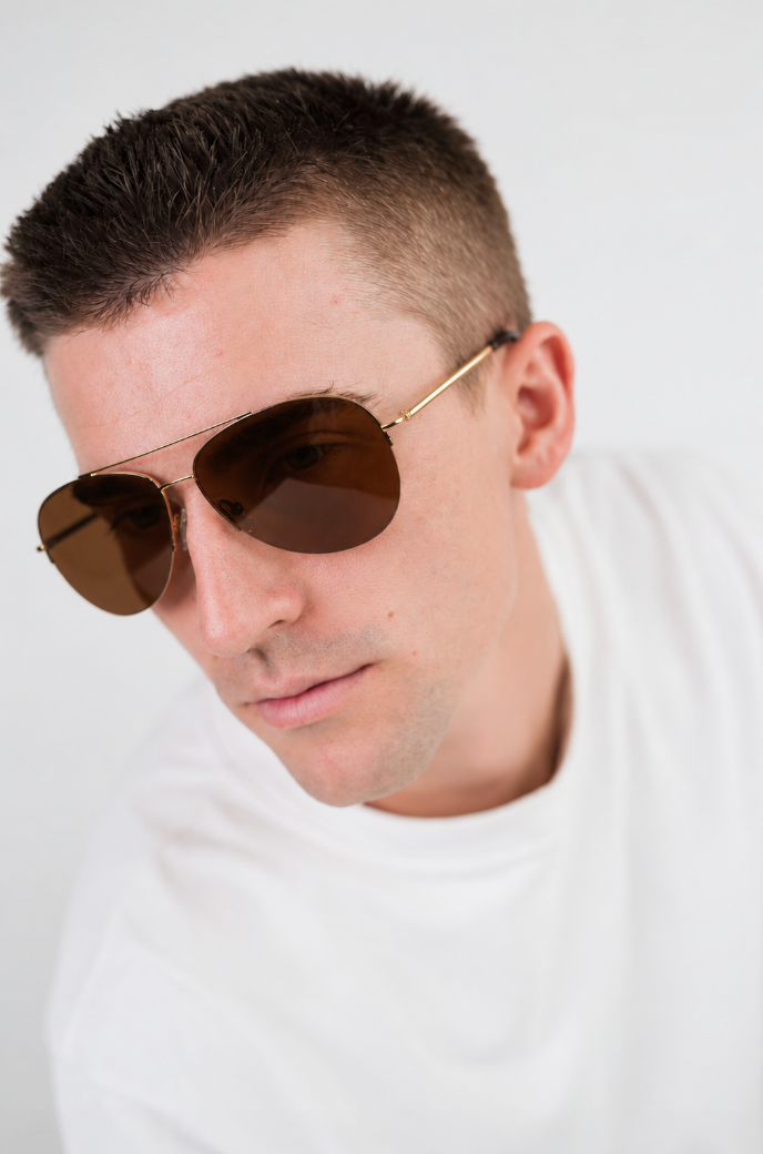 A close-up shot of a man wearing aviator sunglasses with a gold metal frame and dark brown lenses, paired with a white t-shirt against a plain white background.
