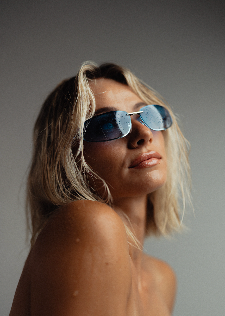 A close-up portrait of a woman with blonde hair wearing blue-tinted aviator sunglasses against a neutral gray background.