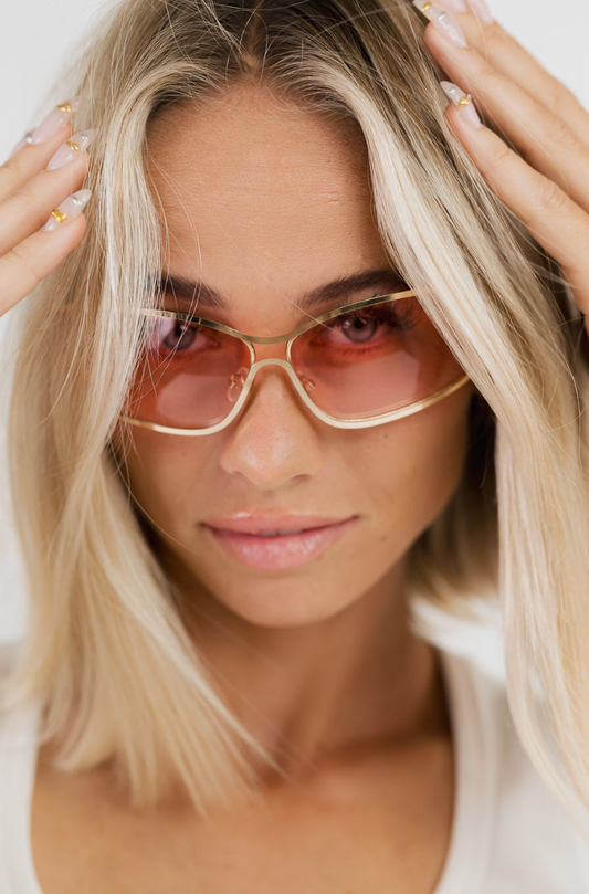 A close-up of a blonde woman wearing oversized square sunglasses with a gold metal frame and translucent pink lenses, posing with her hands in her hair against a clean white background.
