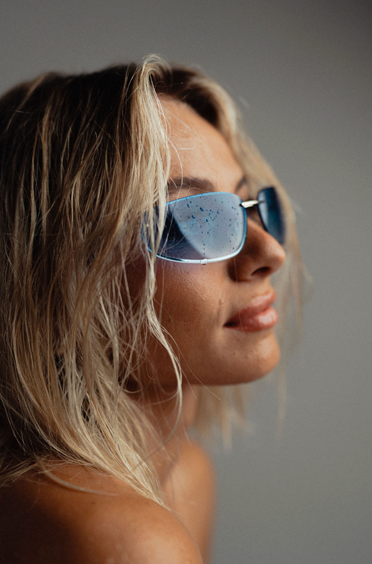 A close-up side profile of a woman with blonde hair wearing rectangular sunglasses with blue-tinted lenses against a plain gray background.