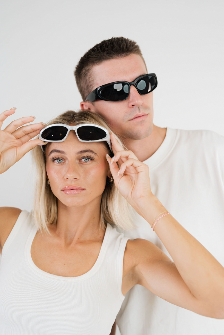 A man and a woman wearing minimalist black sunglasses with white and dark frames, posed against a clean white background.