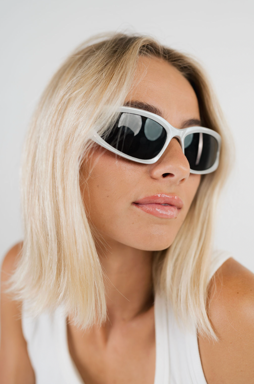 A close-up of a woman with blonde hair wearing white cat-eye sunglasses with dark lenses and a white tank top, set against a plain light background.
