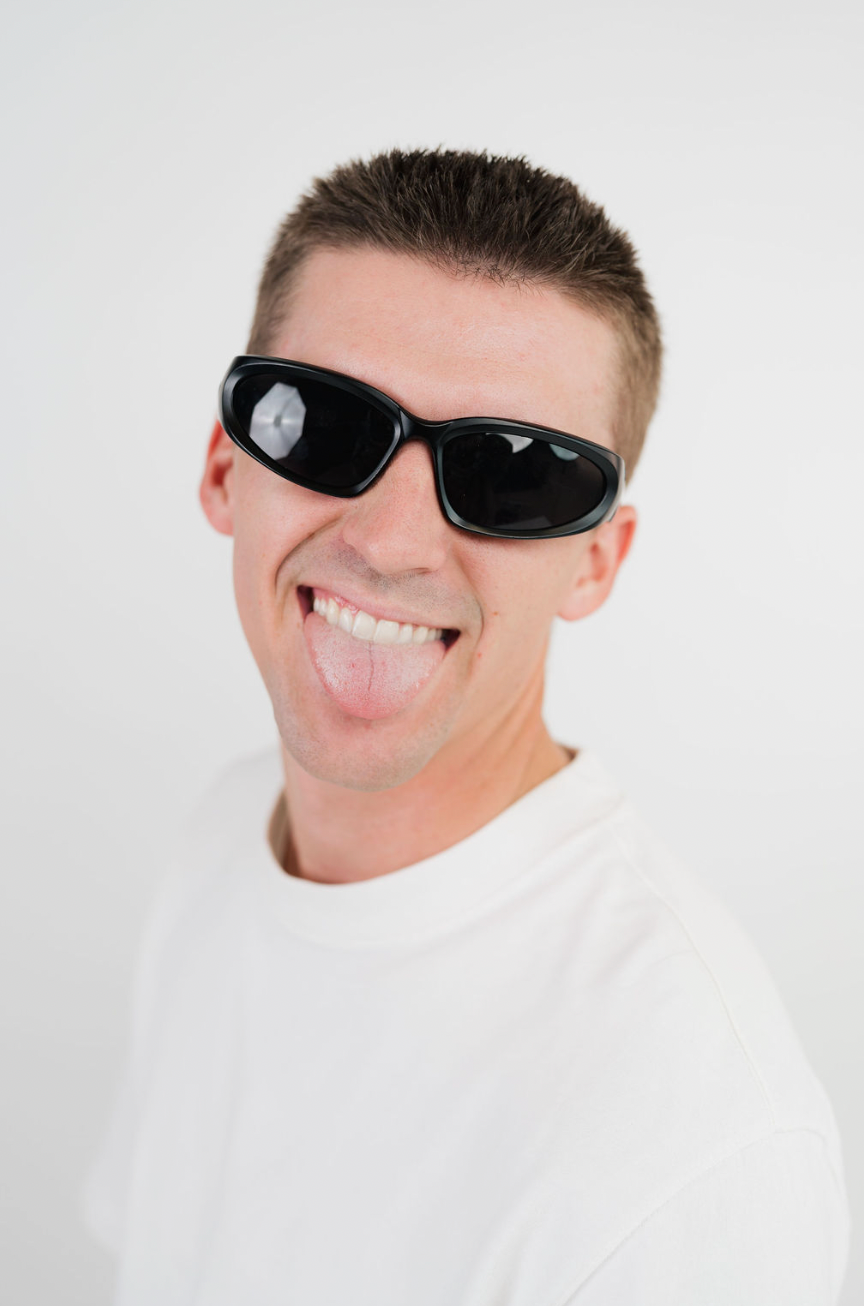 A man wearing black sunglasses with a circular reflective mirror on the left lens, sticking his tongue out against a plain white background.