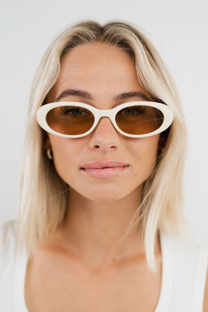 A close-up of a woman with blonde hair wearing white cat-eye sunglasses with amber-brown tinted lenses, set against a clean, light gray background.