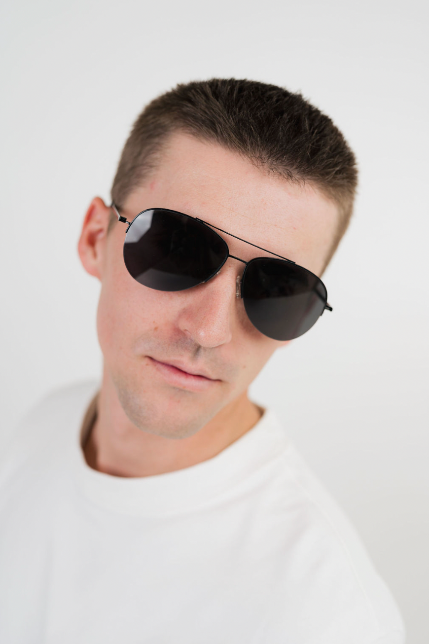 A man wearing classic aviator sunglasses with dark lenses and thin metal frames, photographed against a clean white background.