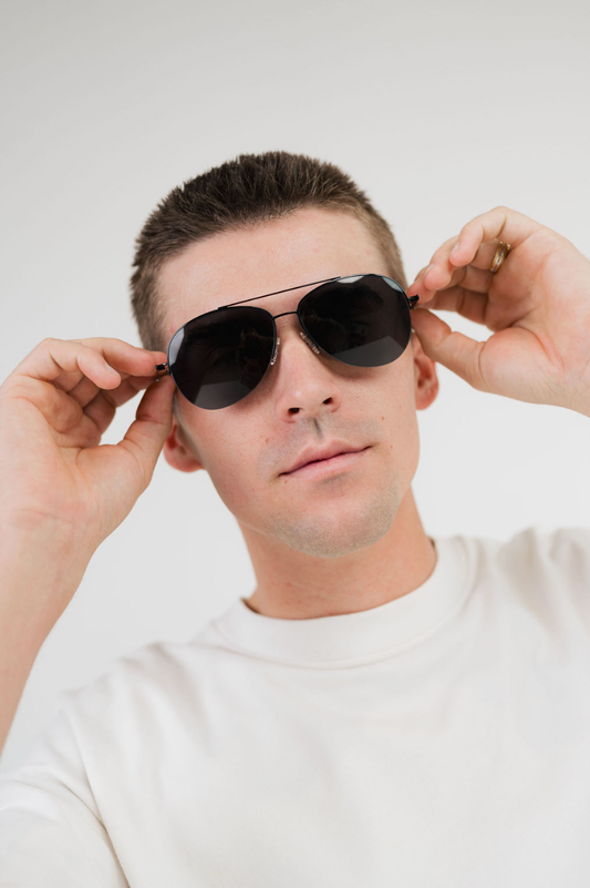 A man wearing aviator-style sunglasses with dark lenses and thin metal frames, holding the sides of the glasses with both hands against a plain light gray background.