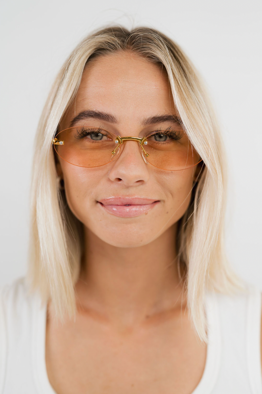 A close-up portrait of a woman with blonde hair wearing thin gold-framed sunglasses with tinted orange lenses, set against a clean white background.