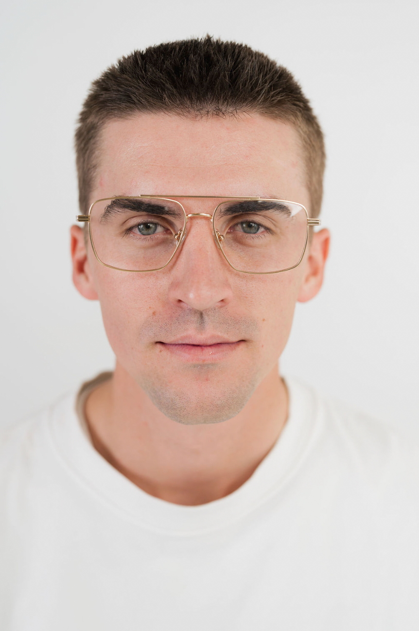 A man with short brown hair wearing gold-framed aviator-style eyeglasses and a white crew-neck t-shirt, photographed against a clean white background.