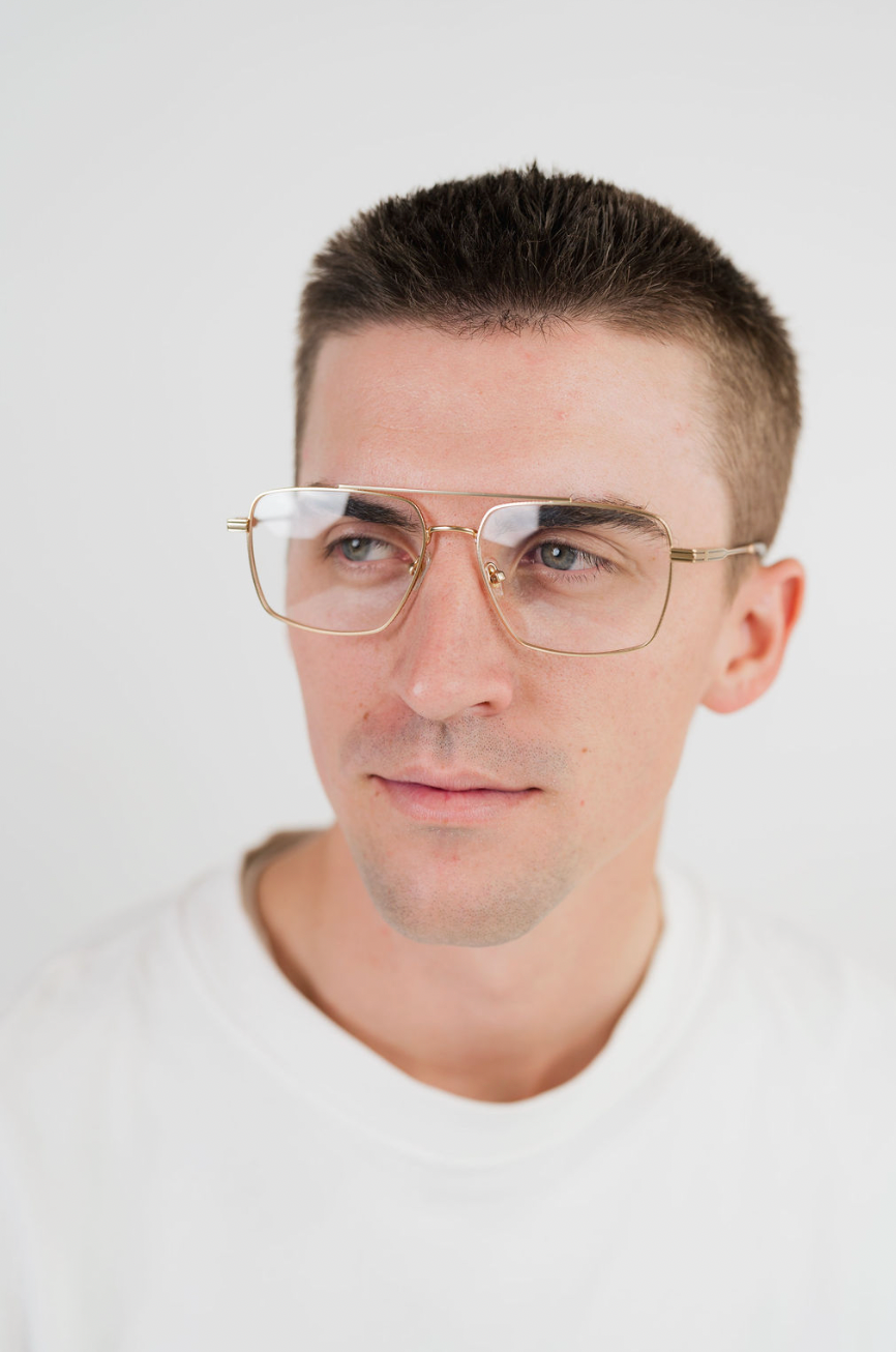 A close-up portrait of a man wearing square aviator-style glasses with a thin gold metal frame and clear lenses, set against a plain light gray background.