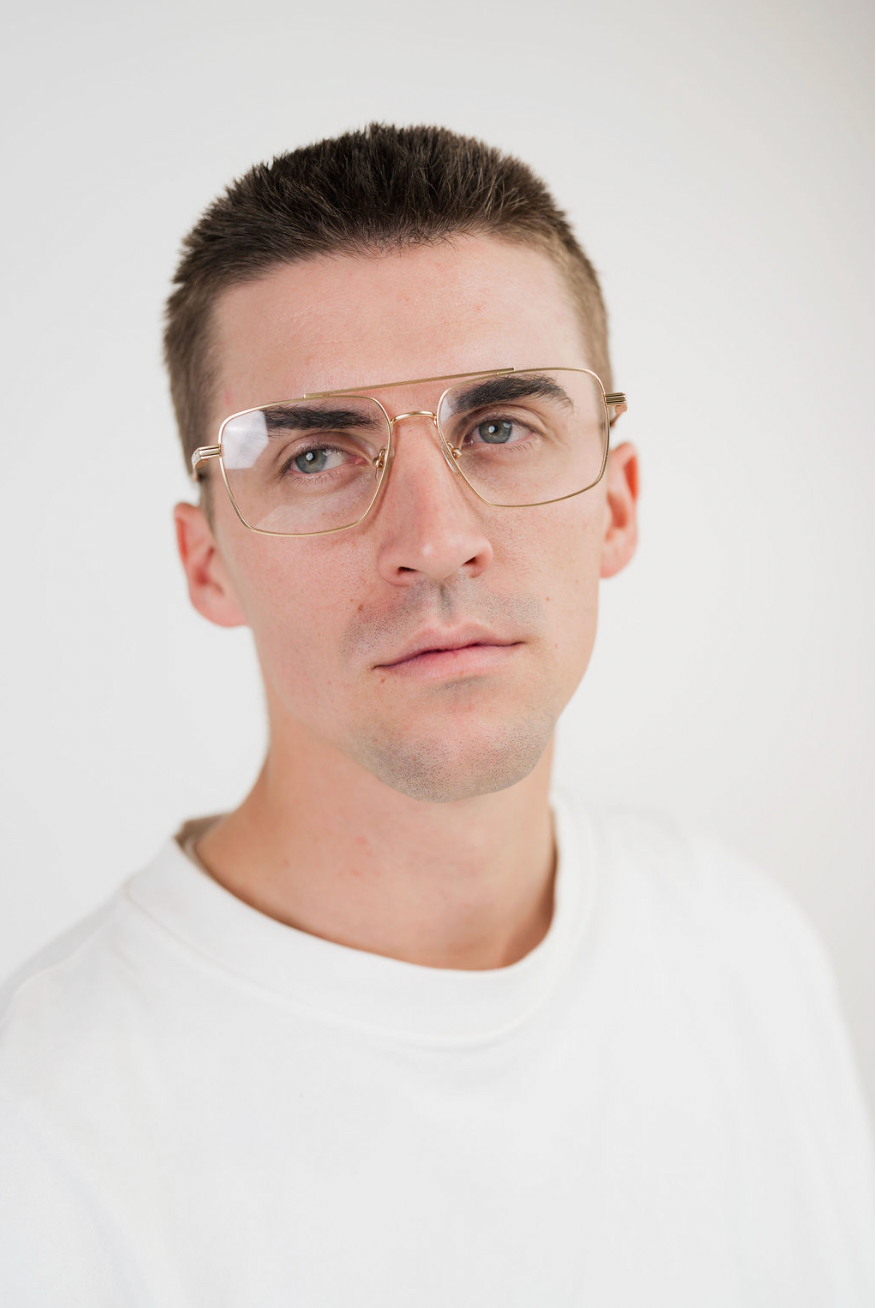 A man with short brown hair wearing gold-framed aviator-style eyeglasses and a white crew-neck t-shirt, photographed against a clean white background.