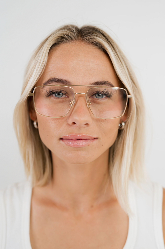 A close-up portrait of a blonde woman wearing aviator-style eyeglasses with a thin gold metal frame and clear lenses, set against a plain light gray background.