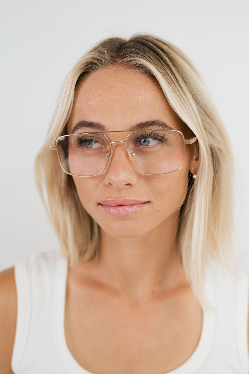 A close-up portrait of a blonde woman wearing oversized square aviator-style glasses with a thin gold metal frame and clear lenses, paired with a white tank top against a plain light background.