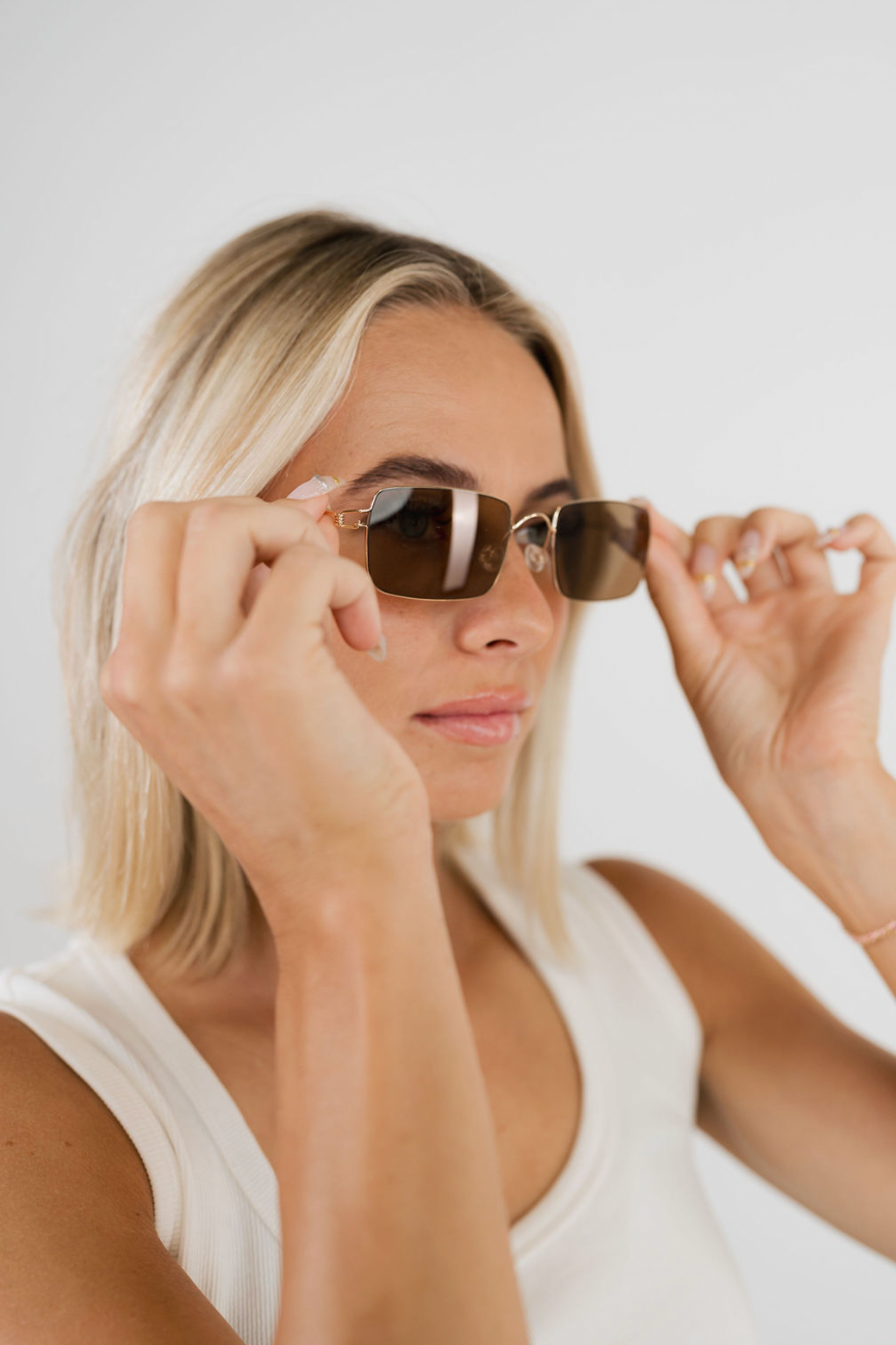 A blonde woman wearing square sunglasses with gold-toned metal frames and dark brown lenses, holding them in place with both hands against a clean white background.