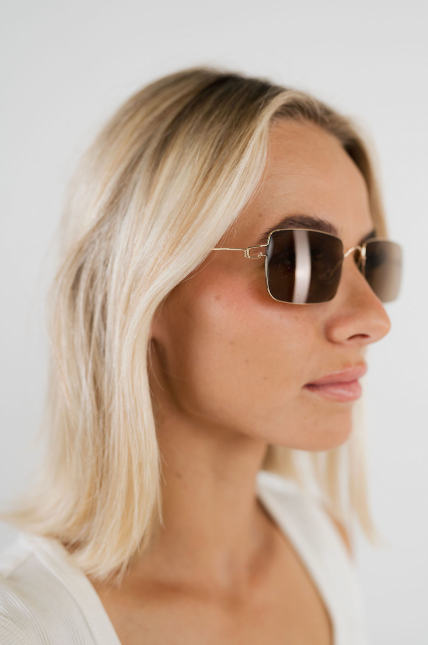 A close-up side profile of a blonde woman wearing slim square sunglasses with a thin gold metal frame and dark brown tinted lenses, set against a clean white background.