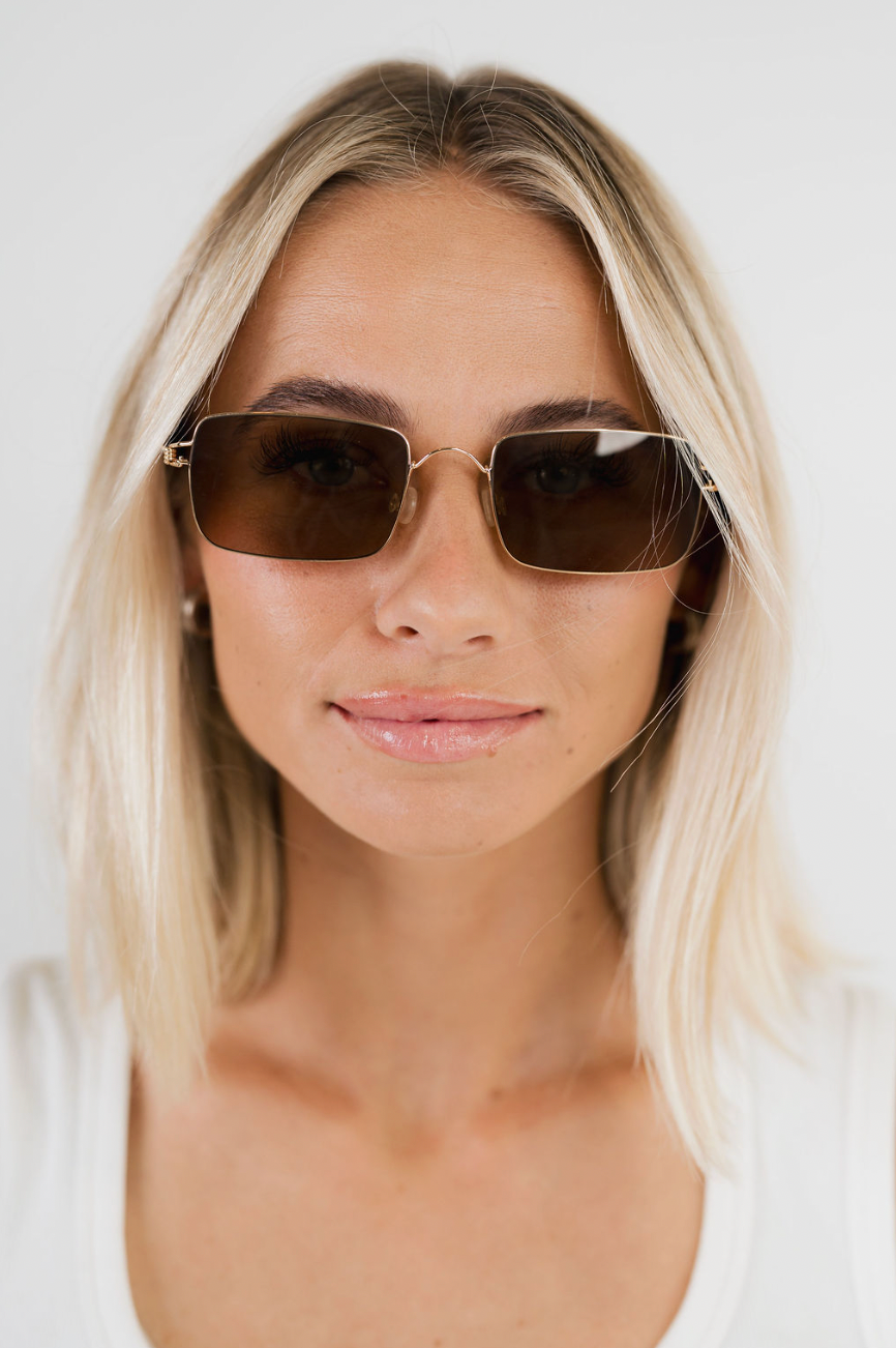 A close-up shot of a blonde woman wearing square-frame sunglasses with dark brown lenses and a thin gold metal frame, set against a plain white background.