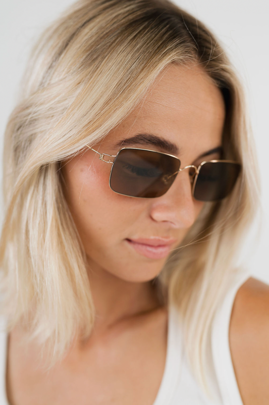 A close-up lifestyle shot of a woman with blonde hair wearing gold-framed square sunglasses with dark brown lenses.