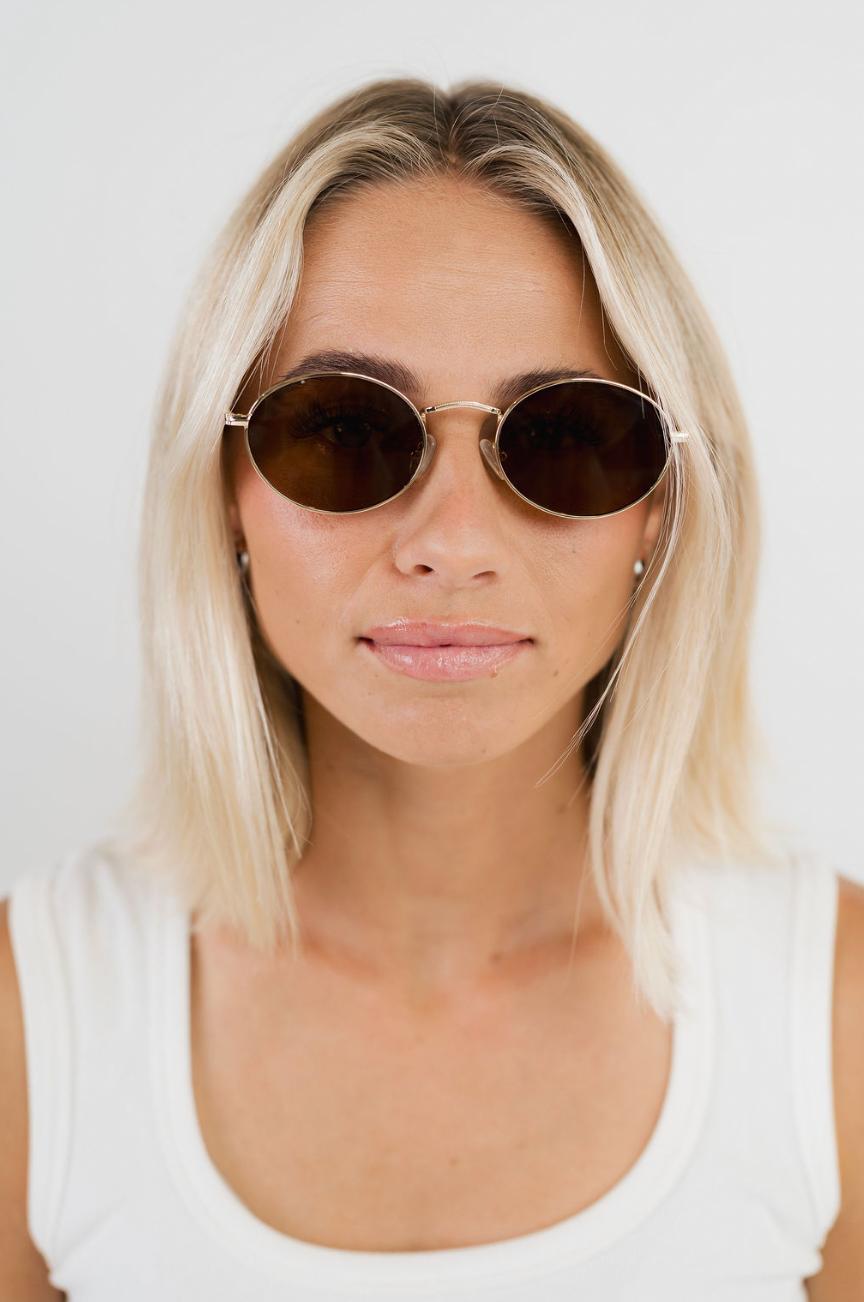 A woman with blonde hair wearing round gold-framed sunglasses with dark brown lenses and a white tank top, photographed against a plain white background.