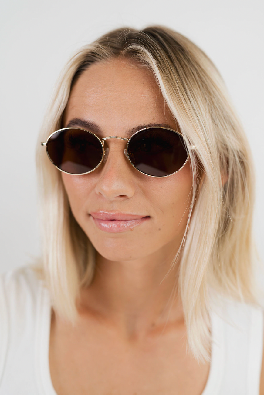 A close-up of a woman with blonde hair wearing round gold-framed sunglasses with dark lenses, set against a plain light background.