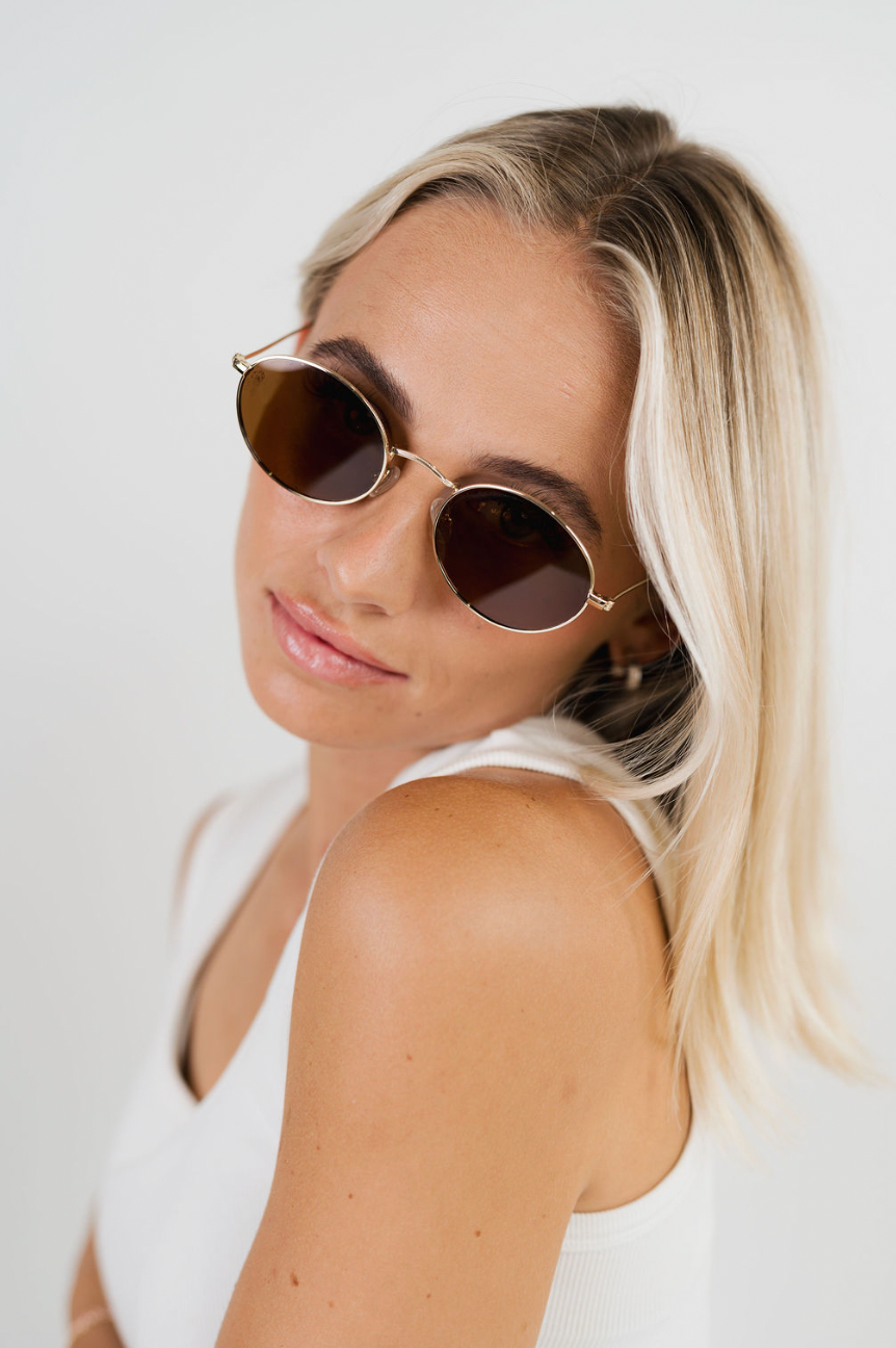 A blonde woman wearing round gold-framed sunglasses with dark brown lenses, posing against a clean white background.