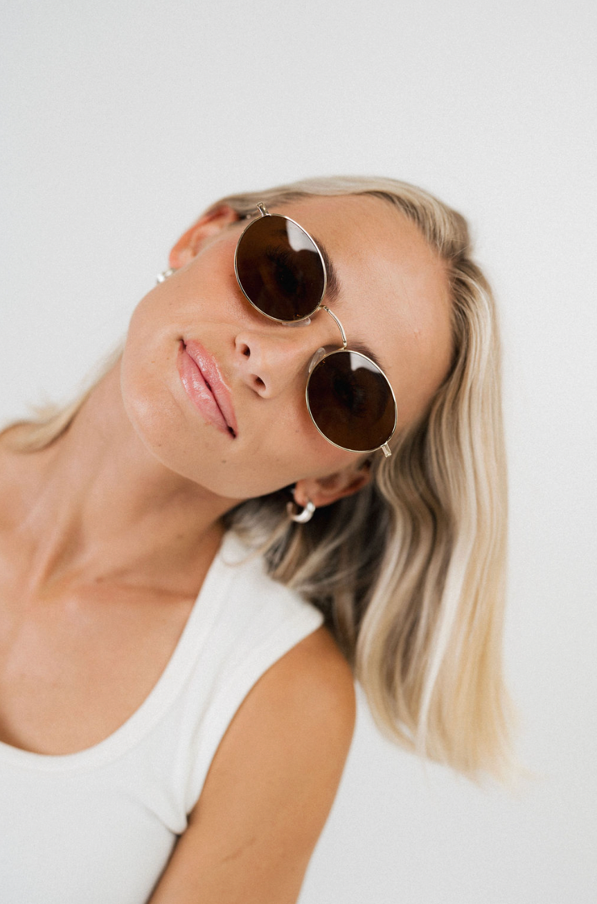 A close-up lifestyle shot of a blonde woman wearing round sunglasses with dark brown lenses and thin gold metal frames, paired with a white tank top against a plain light background.