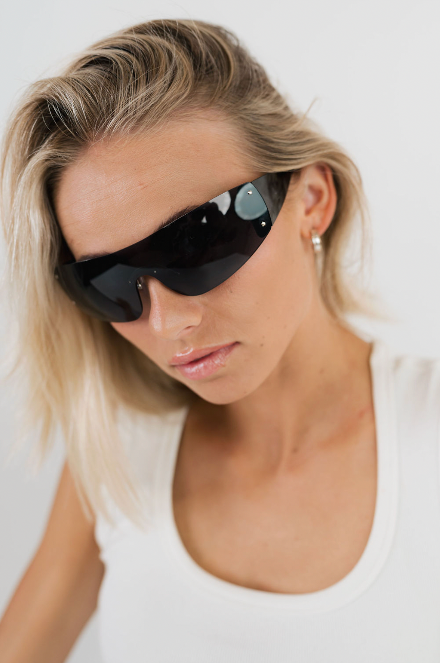 A close-up of a blonde woman wearing black wrap-around shield sunglasses and a white tank top against a plain light background.