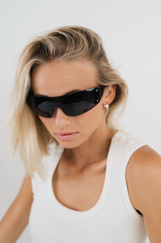 A woman with blonde hair wearing black shield-style sunglasses and a white tank top, photographed against a plain light background.