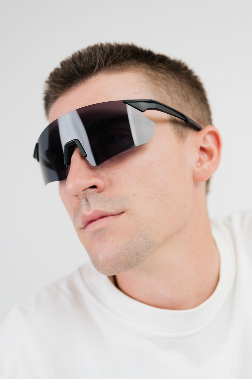 A close-up of a man wearing black wrap-around shield sunglasses with a dark reflective lens, set against a plain light gray background.