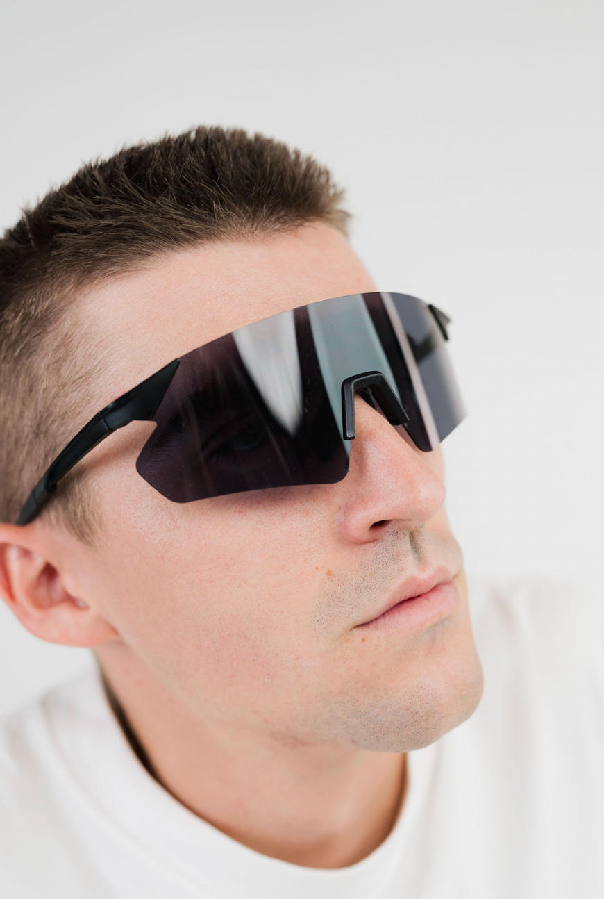 A close-up of a man wearing futuristic wraparound shield-style sunglasses with a dark reflective lens and black frame, set against a plain light gray background.
