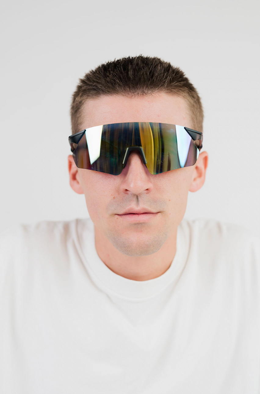 A close-up of a man wearing shield-style sunglasses with a reflective, iridescent mirror finish. The lenses show a blend of green, yellow, and blue reflections.
