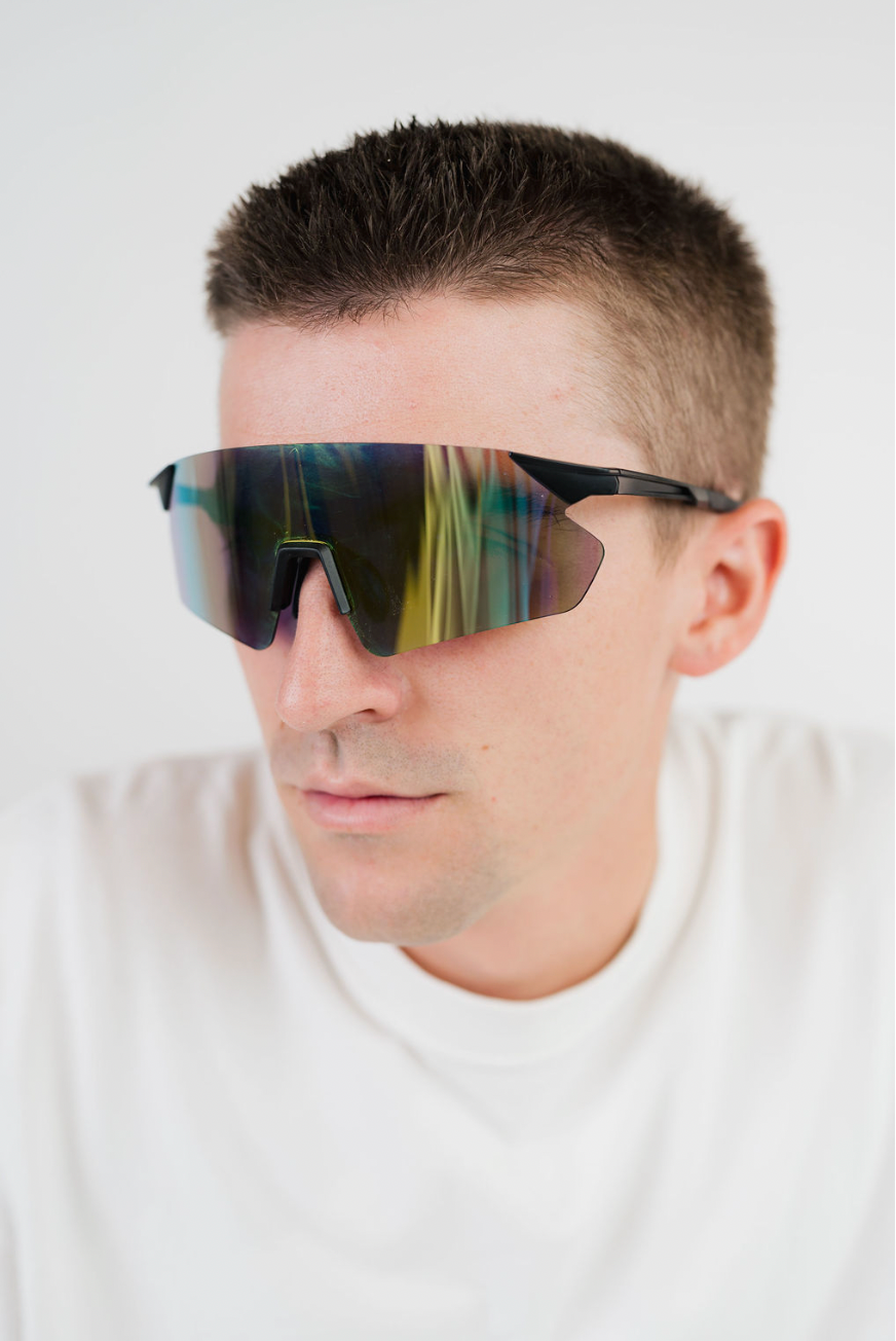 A close-up of a man wearing shield-style sunglasses with a black frame and a reflective dark lens that shows rainbow-like reflections, against a plain light background.