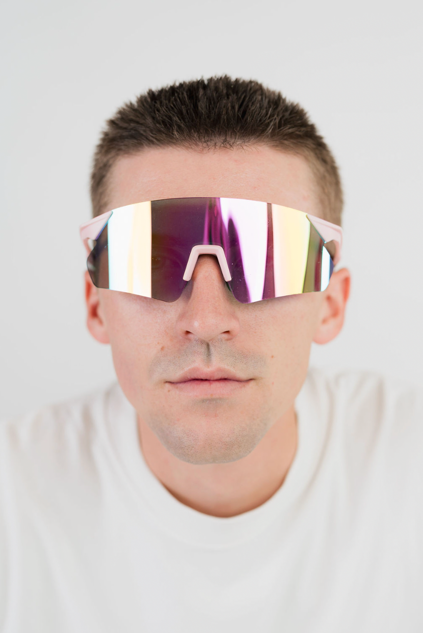 A close-up of a man wearing futuristic wraparound shield-style sunglasses with a reflective iridescent mirror lens and light pink arms, set against a plain light gray background.