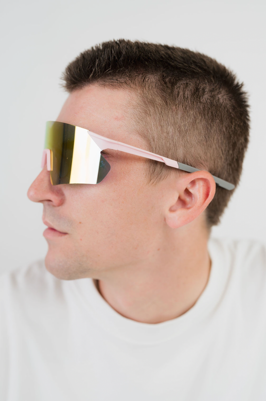 A man in profile wearing futuristic shield-style sunglasses with a reflective gold mirrored lens and light pink arms, against a plain light gray background.