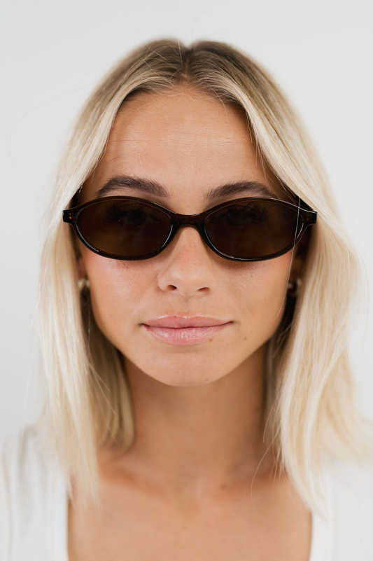 A close-up shot of a woman with blonde hair wearing dark brown cat-eye sunglasses against a plain white background.
