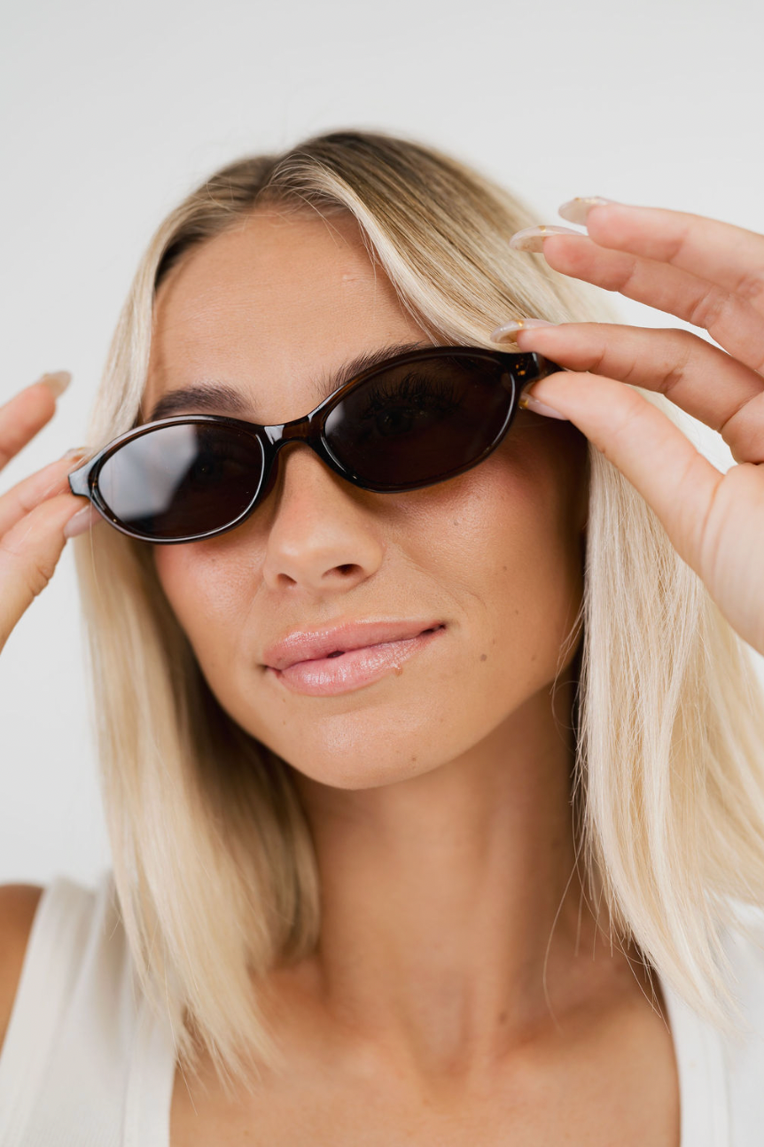 A close-up of a woman with blonde hair wearing black oval sunglasses, holding them in place with both hands against a plain light background.