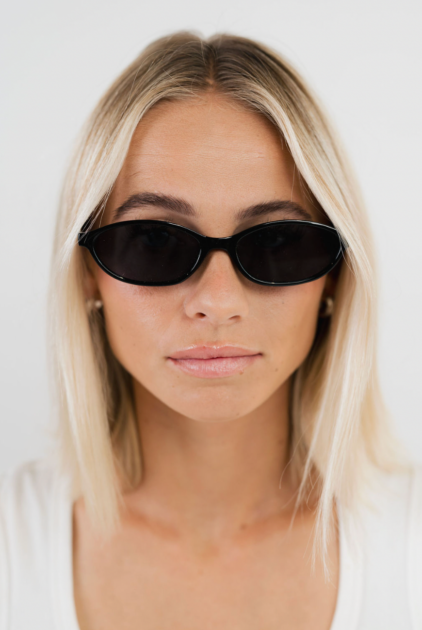 A close-up portrait of a blonde woman wearing black cat-eye sunglasses and a white top against a plain light background.