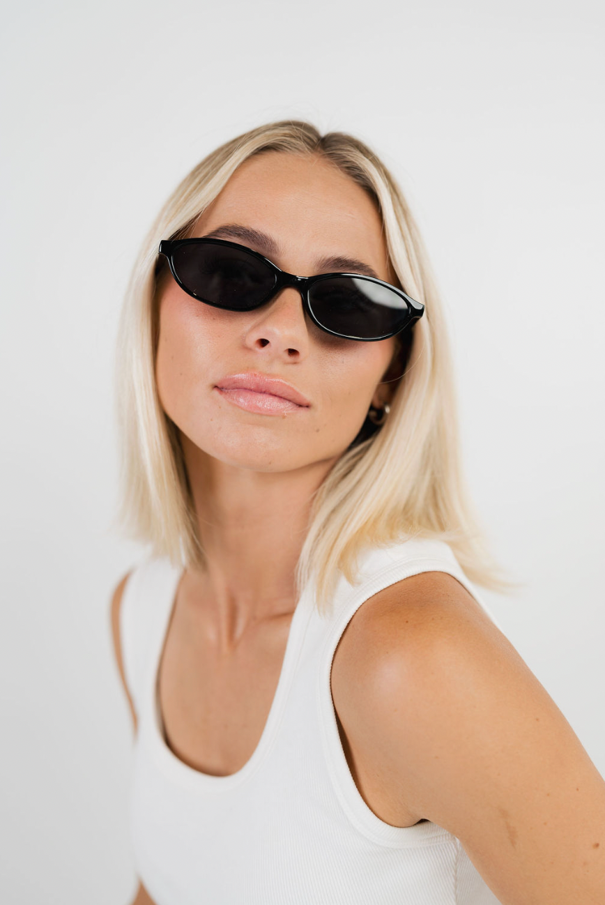 A blonde woman wearing black cat-eye sunglasses and a white tank top, photographed against a clean white background.