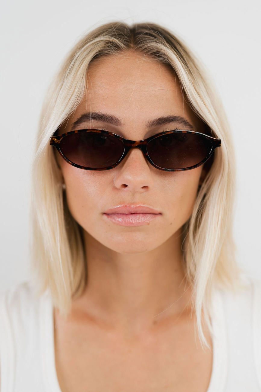 A close-up of a woman with blonde hair wearing dark tortoiseshell cat-eye sunglasses and a white top, set against a plain white background.