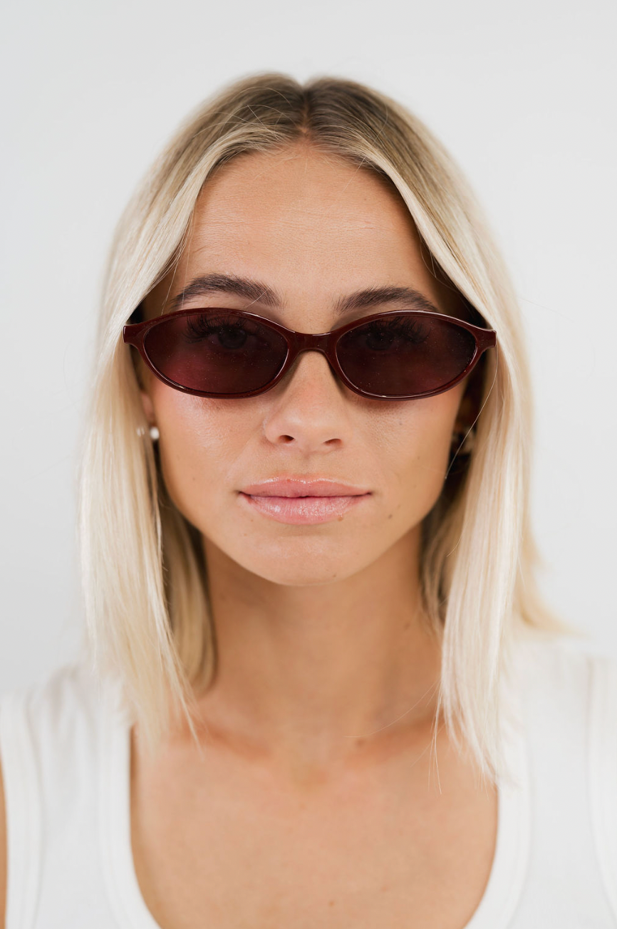A close-up of a woman with blonde hair wearing dark brown cat-eye sunglasses and a white tank top against a plain white background.