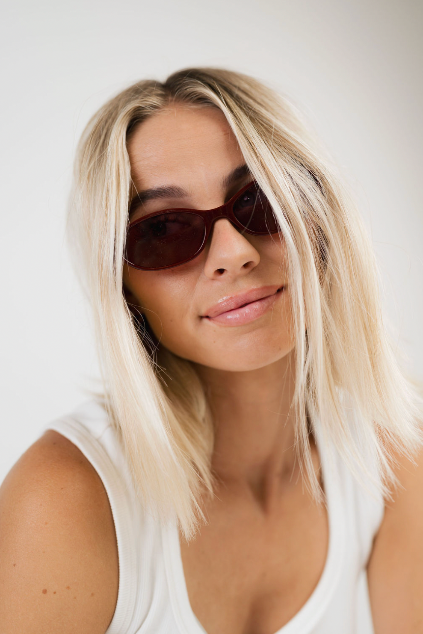 A close-up portrait of a woman with blonde hair wearing dark brown sunglasses and a white tank top against a plain light background.