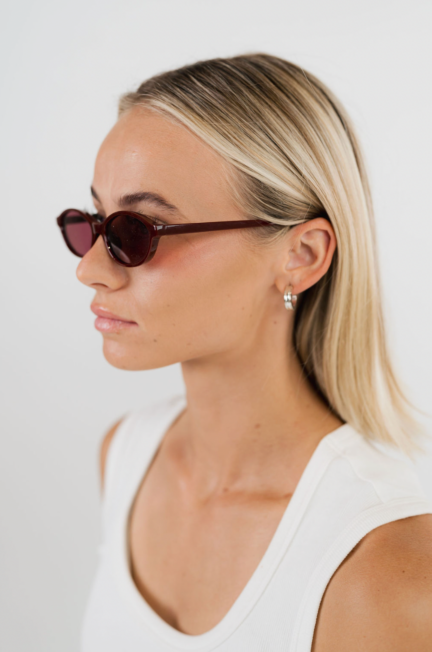 A blonde woman in profile wearing dark brown acetate sunglasses with a small silver hoop earring, against a clean white background.