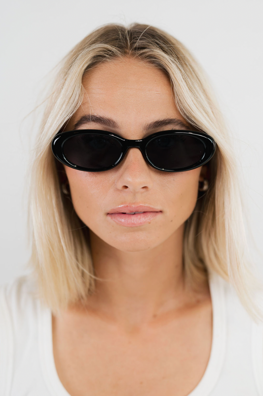 A close-up of a woman with blonde hair wearing black oval sunglasses and a white top against a plain light background.