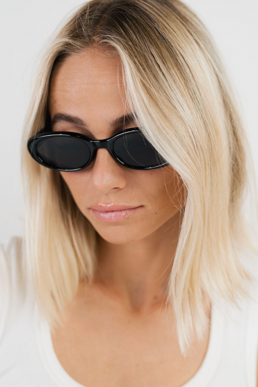 A close-up of a woman with blonde hair wearing black cat-eye sunglasses and a white top against a plain white background.