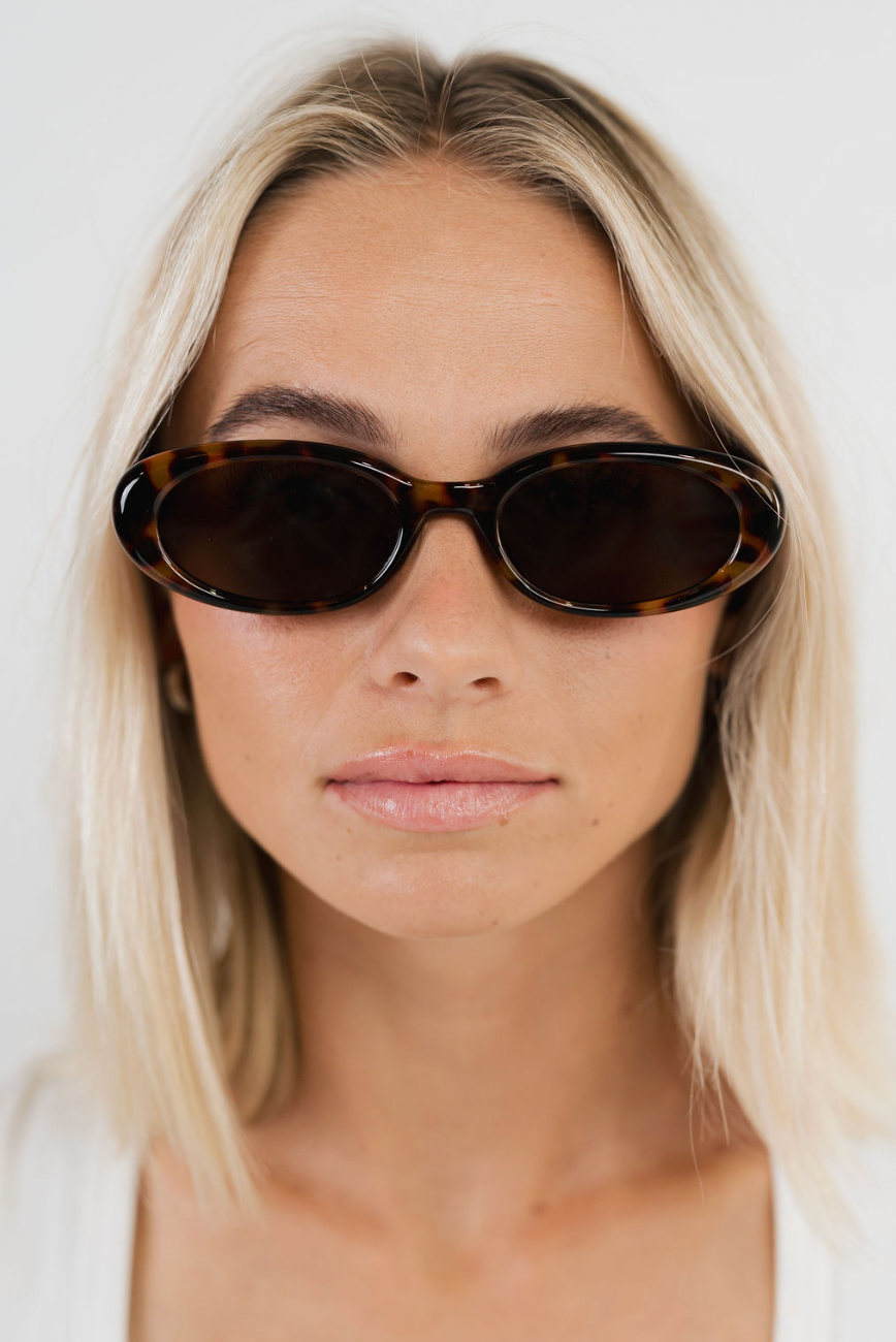 A close-up of a blonde woman wearing dark brown tortoiseshell oval sunglasses against a clean white background.