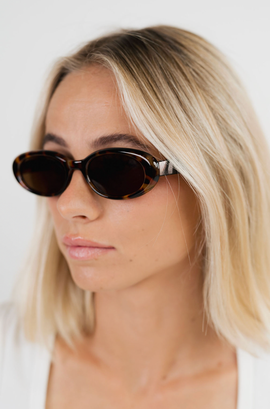 A close-up of a blonde woman wearing dark brown tortoiseshell oval sunglasses with silver metal temples, set against a clean white background.