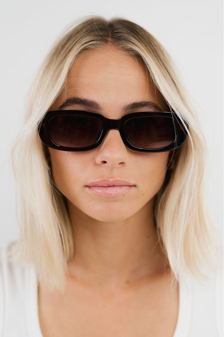 A close-up portrait of a blonde woman wearing black rectangular sunglasses with dark lenses, set against a clean white background.