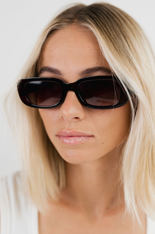 A close-up of a woman with blonde hair wearing black cat-eye sunglasses against a plain white background.